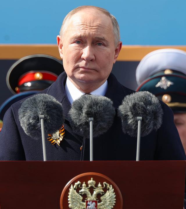 Ein älterer Mann im Anzug steht hinter einem Rednerpult mit einem Adlerwappen und drei Mikrofonen. Hinter ihm sieht man die Mützen von Militärs.