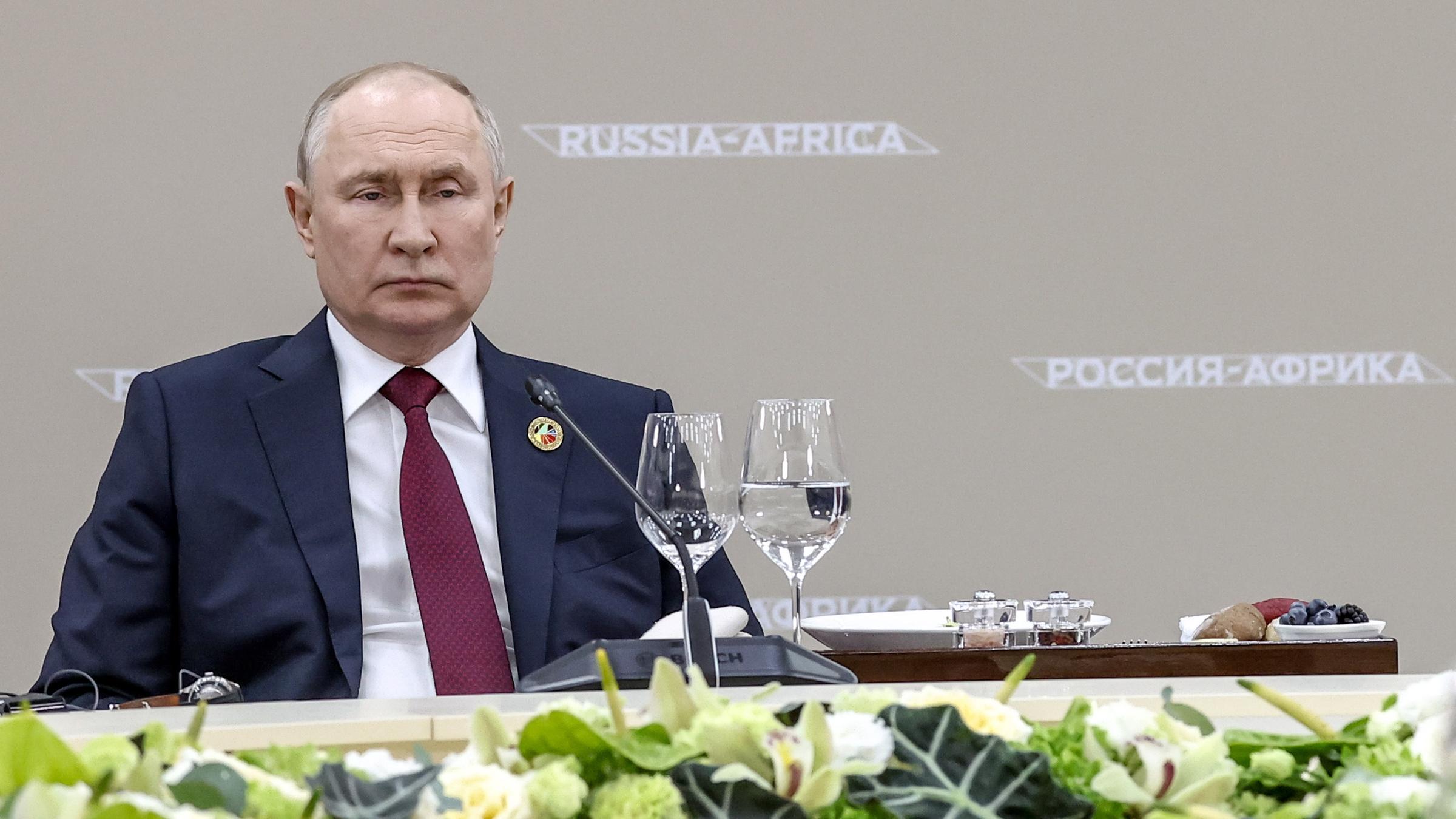 Zu sehen ist Präsident Putin an einem Konferenztisch mit Blumen im Vordergrund.