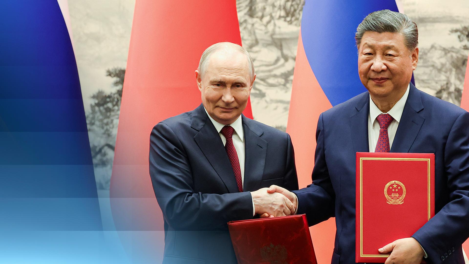 Vladimir Putin bei seinem Besuch in Peking schüttelt Präsident Xi die Hand, während beide in rotem Leder gebundene Umschläge halten. Hinter ihnen sind die Nationalflaggen von China und Russland zu sehen.
