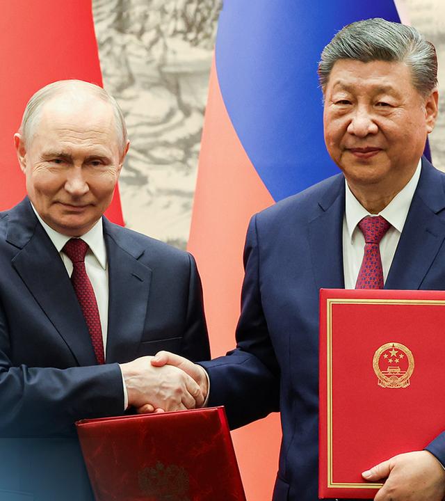 Vladimir Putin bei seinem Besuch in Peking schüttelt Präsident Xi die Hand, während beide in rotem Leder gebundene Umschläge halten. Hinter ihnen sind die Nationalflaggen von China und Russland zu sehen.
