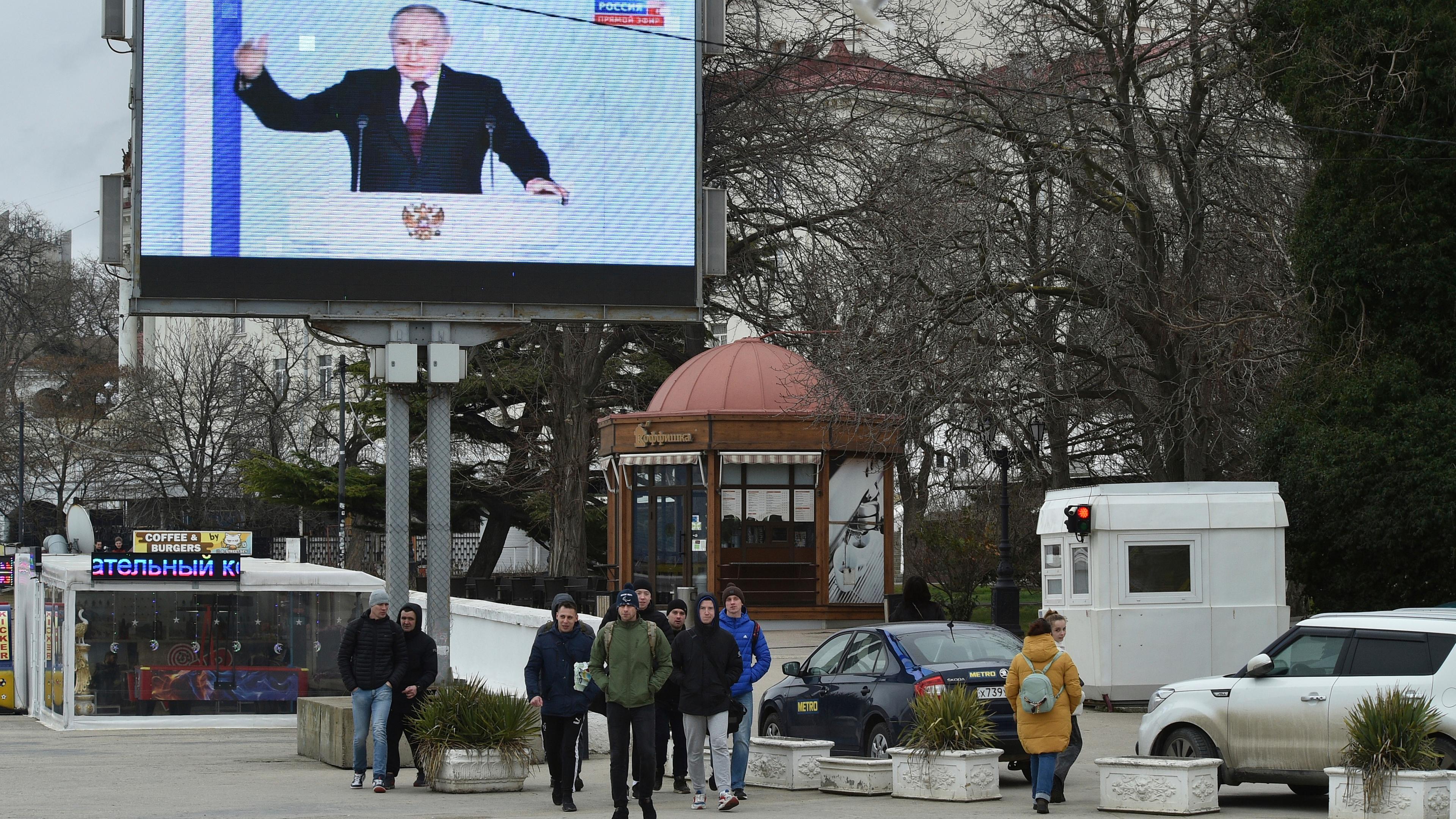 Archiv: Menschen gehen vor einem Fernsehbildschirm, der den russischen Präsidenten Putin während seiner jährlichen Rede zur Lage der Nation in Sewastopol auf der Krim zeigt.  am 21.02.2023