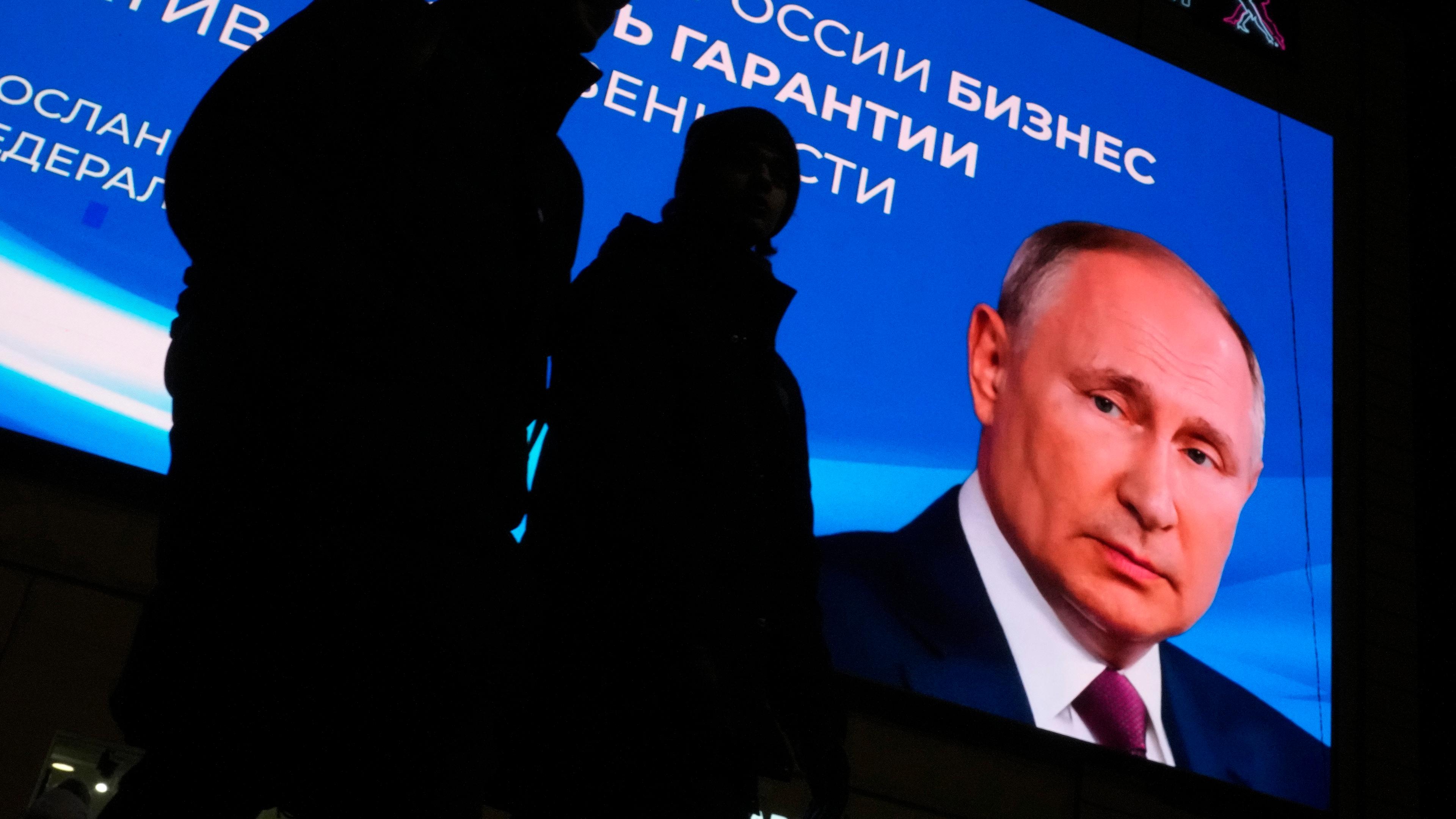Beleuchtetes Wahlplakat von Putin im Hintergrund. Die Silhouetten von zwei Passanten im Vordergrund.