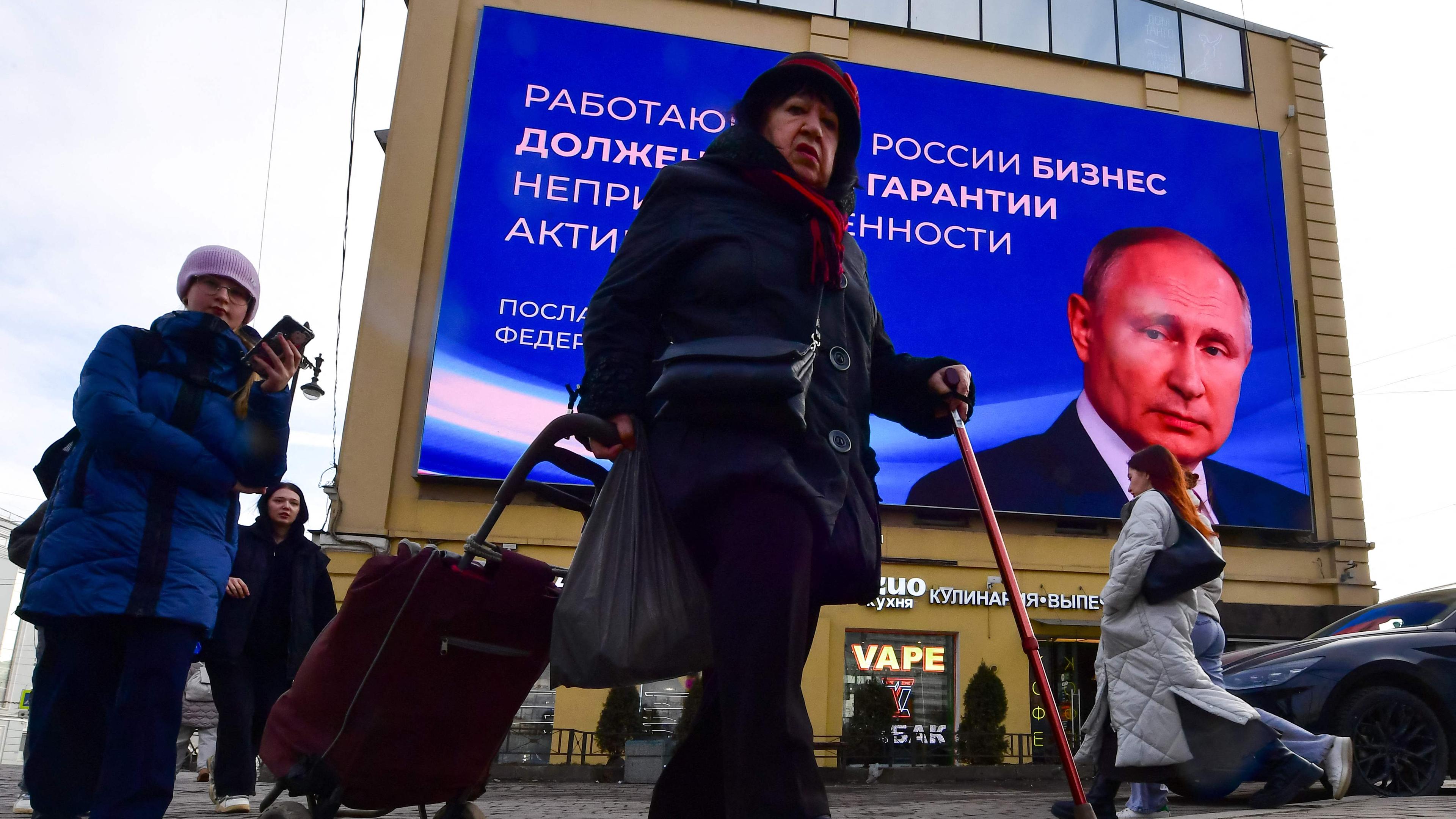 Passanten in der Fußgängerzone in Sankt Petersburg. Im Hintergrund ein großes digitales Werbeplakat von Wladimir Putin.