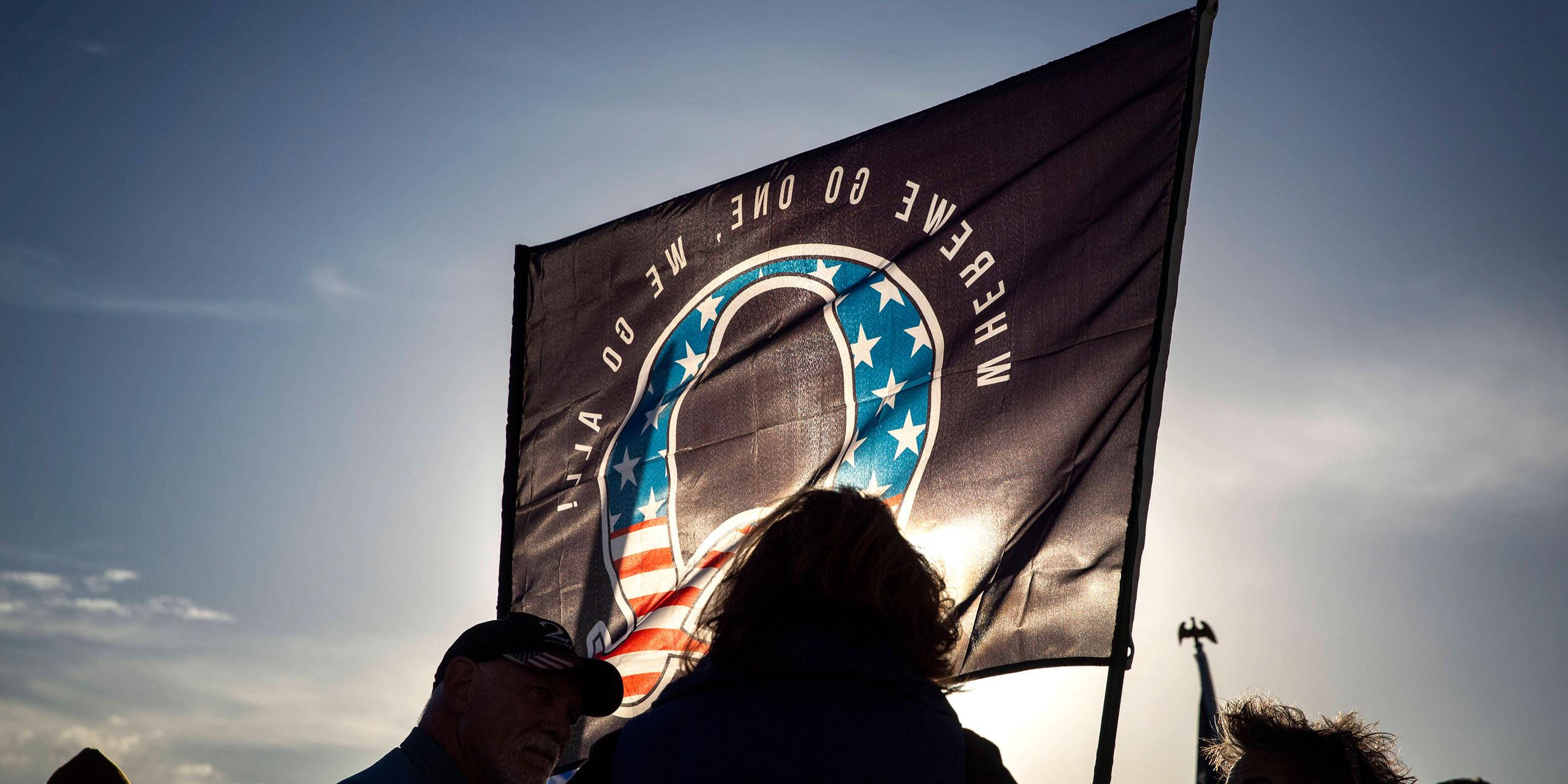 Vor einer von der untergehenden Sonne angeleuchteten Flagge mit einerm großen Q in den Farben der amerikanischen Flagge, sind die Silhouetten mehrerer Personen dunkel zu sehen.