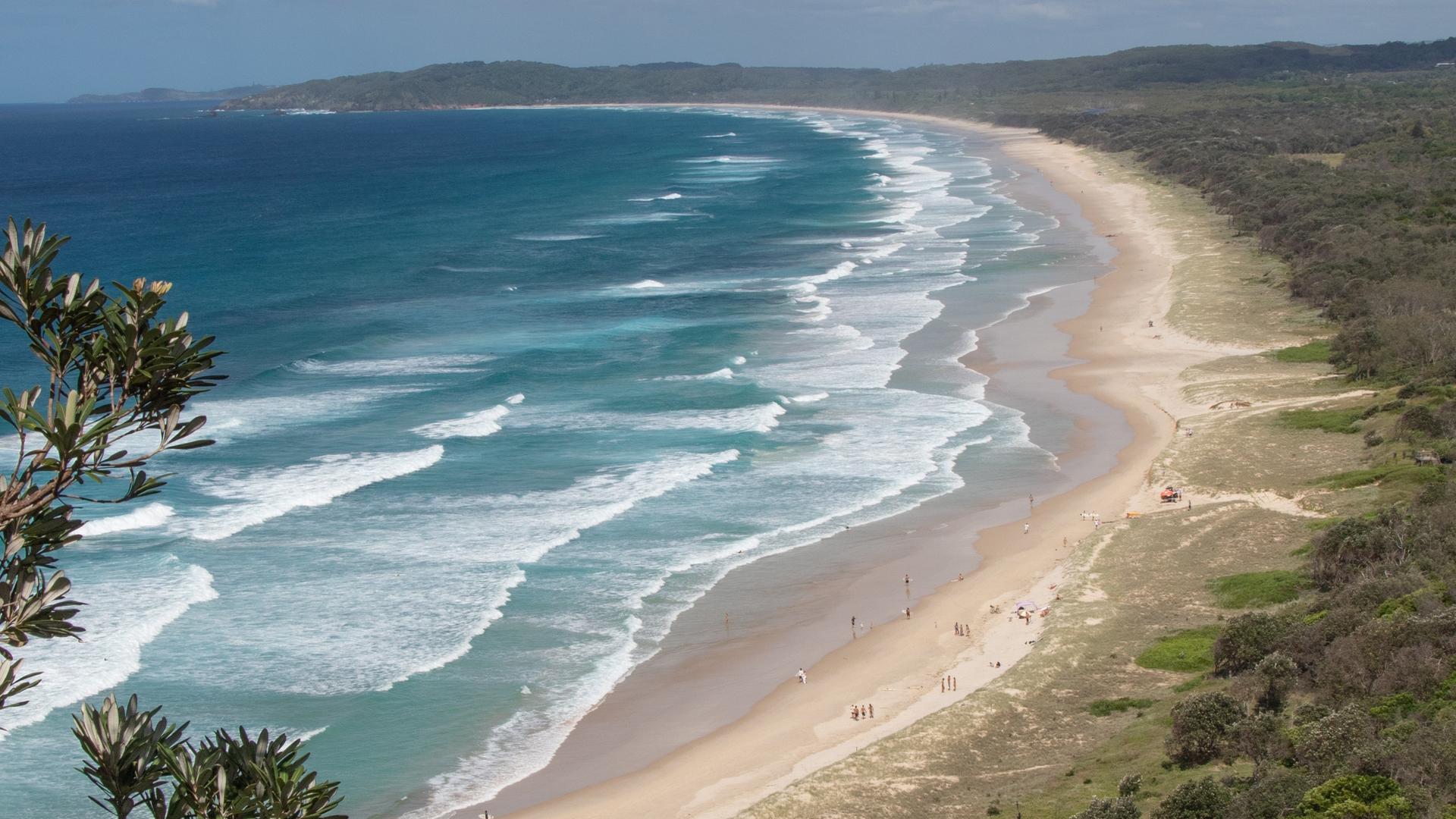 Panoramaaufnahme eines weitläufigen Sandstrandes der in ein dichtes Waldgebiet übergeht. Am blauem Himmel hängen einige Wolken.