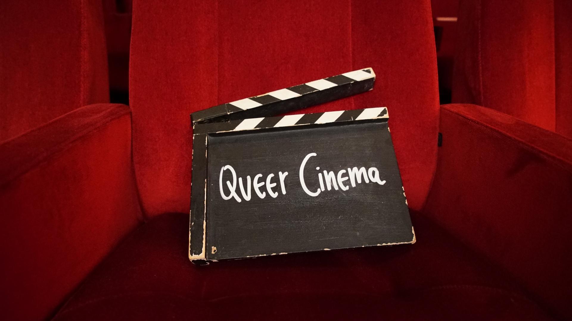 Eine Filmklappe mit der Aufschrift „Queer Cinema“ steht in einer Seitenische eines schmalen Ganges mit schwarzem Boden und roten Wänden.