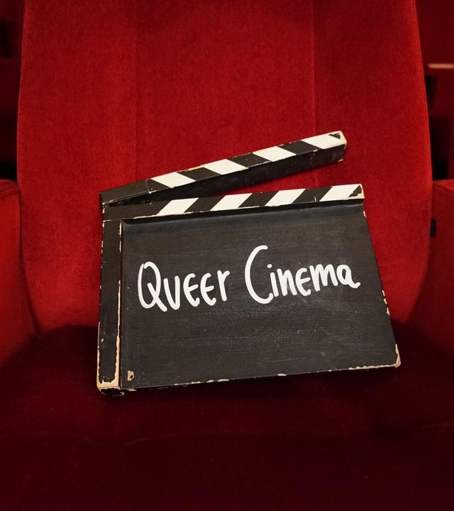 Eine Filmklappe mit der Aufschrift „Queer Cinema“ steht in einer Seitenische eines schmalen Ganges mit schwarzem Boden und roten Wänden.