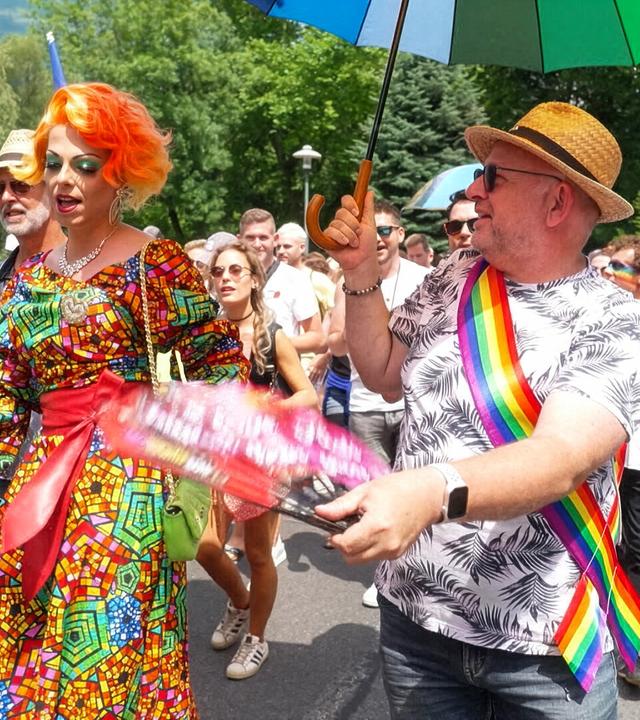 Das Bild zeigt eine Szene von einer Pride Parade in Innsbruck, Österreich. Im Vordergrund sind mehrere Teilnehmer zu sehen, die fröhlich marschieren. Eine Person trägt ein auffälliges, buntes Kleid mit geometrischem Muster sowie eine orangefarbene Perücke und hat eine bewundernde Gestik. Neben ihr ist ein weiterer Teilnehmer, der ein Hemd mit tropischem Muster trägt und ein Regenbogen-Sternenbanner um die Schulter geworfen hat.   Der Mann hält einen grünen Regenschirm und hat eine fröhliche Miene. Im Hintergrund sind eine große Menschenmenge zu sehen, die ebenfalls feierlich und ausgelassen agiert. Viele tragen bunte Accessoires, wie Regenbogenflaggen oder geschmückte Kleidung. Die Straße ist gesäumt von Bäumen, die eine grüne Umgebung bieten. Die Atmosphäre wirkt festlich und voller Freude, was die Botschaft von Toleranz und Diversität unterstreicht. Der gesamte Anlass vermittelt ein Gefühl von Gemeinschaft und Unterstützung für die LGBTIQ+-Bewegung.