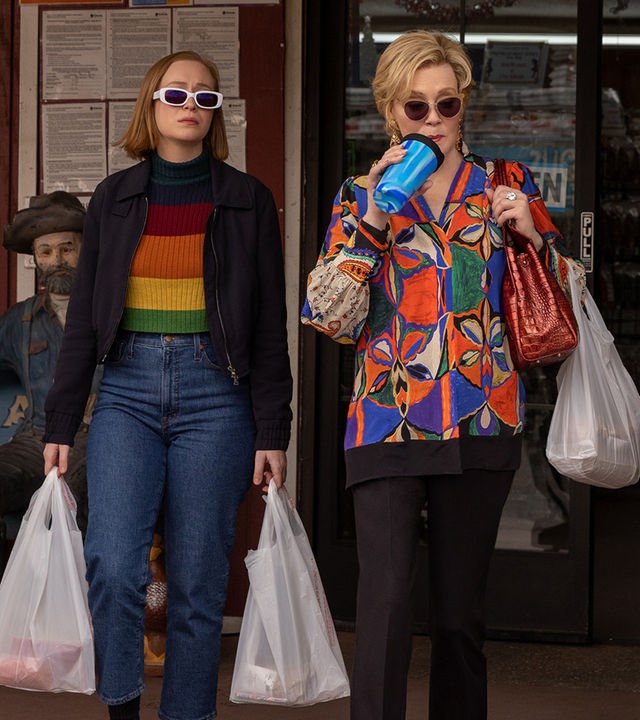 "Hacks - Quid pro quo": (Hannah Einbinder) und Deborah (Jean Smart) tragen Sonnenbrillen und treten mit Einkaufstaschen aus einem Geschäft, während Deborah aus einem Trinkbecher mit Strohhalm trinkt. Neben dem Eingang stehen eine Statue eines beladenen Esels und eine Werbetafel mit Angeboten für verschiedene Fleischsorten.