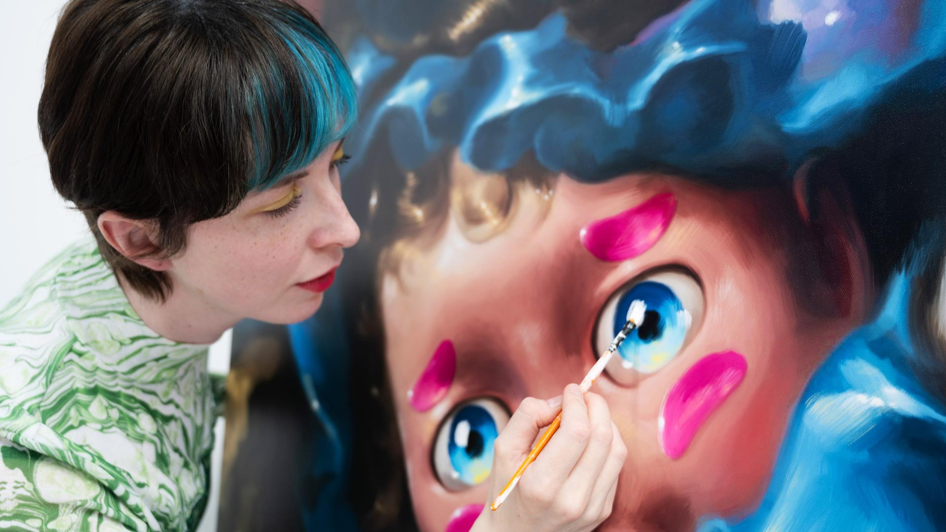 Frau mit kurzen Haaren und blauer Strähne malt mit Pinsel an einem großen puppenartigen Gesicht auf Leinwand.