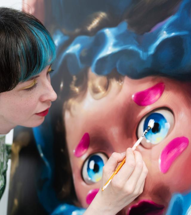 Frau mit kurzen Haaren und blauer Strähne malt mit Pinsel an einem großen puppenartigen Gesicht auf Leinwand.