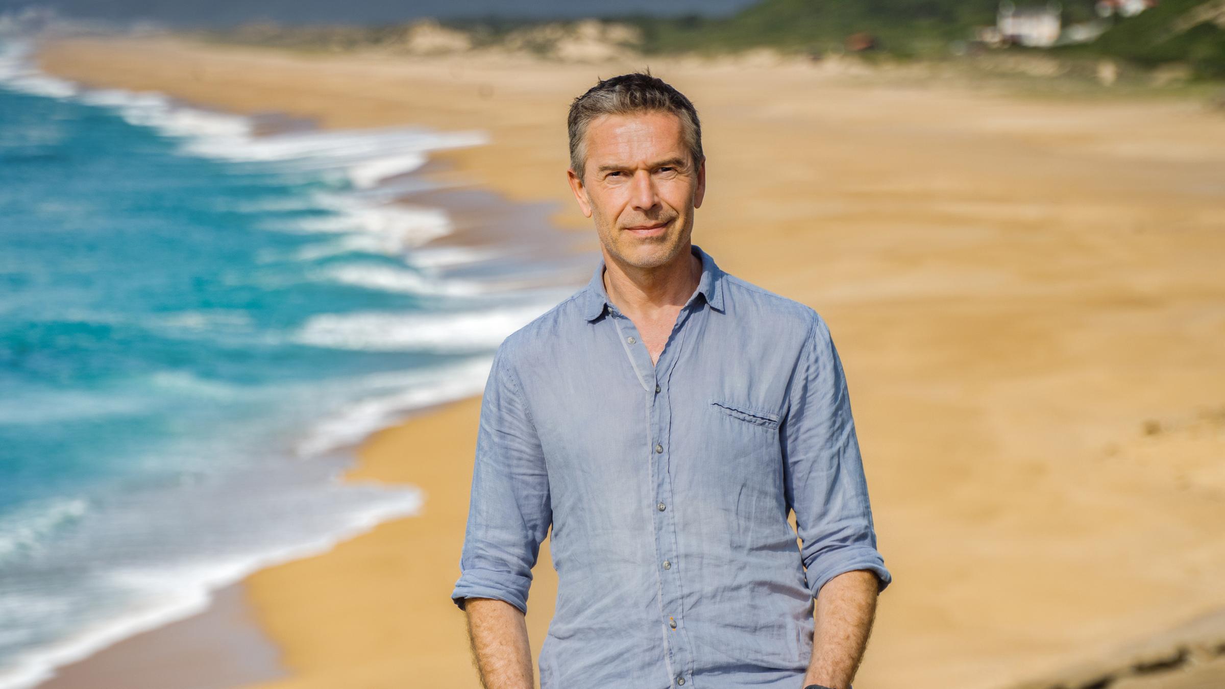 Dirk Steffens steht auf einer Klippe hoch über einem langen Sandstrand.