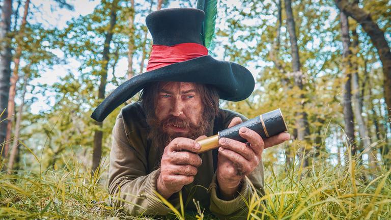 "Der Räuber Hotzenplotz": Der Räuber Hotzenplotz (Nicholas Ofczarek) liegt im Wald auf der Lauer. Sein Blick liegt in der Ferne. Sein Gesichtsausdruck wirkt entschlossen.