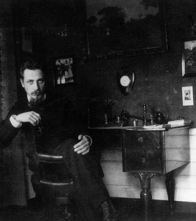 Historisches Schwarz-weiß-Foto von einem jungen Mann mit Bart, der an einem Schreibtisch sitzt und sich zum Betrachter umdreht.