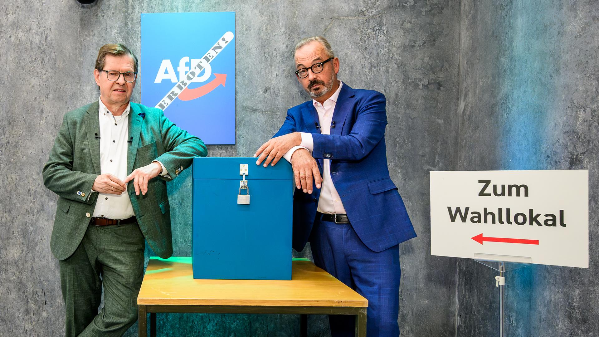 Ralf Stegner und Jan Fleischhauer lehnen an einem Tisch mit einer blauen, durch ein Vorhängeschloss gesicherten Wahlurne. 