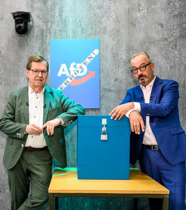 Ralf Stegner und Jan Fleischhauer lehnen an einem Tisch mit einer blauen, durch ein Vorhängeschloss gesicherten Wahlurne. 