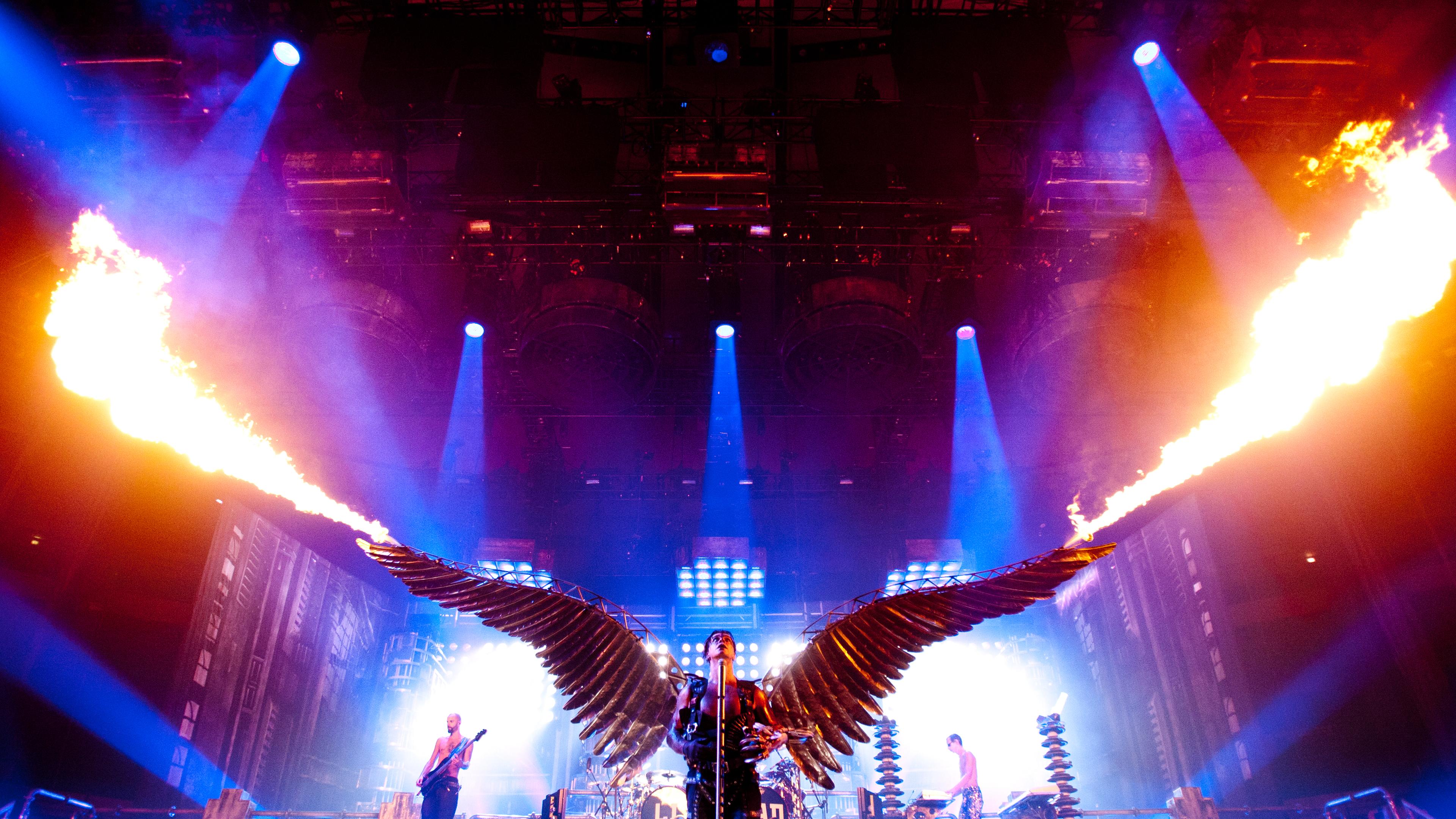 Der Sänger von Rammstein Till Lindemann steht mit großen goldenen Flügeln am Rücken, aus denen Feuer kommt. Die Bühne ist blau beleuchtet.
