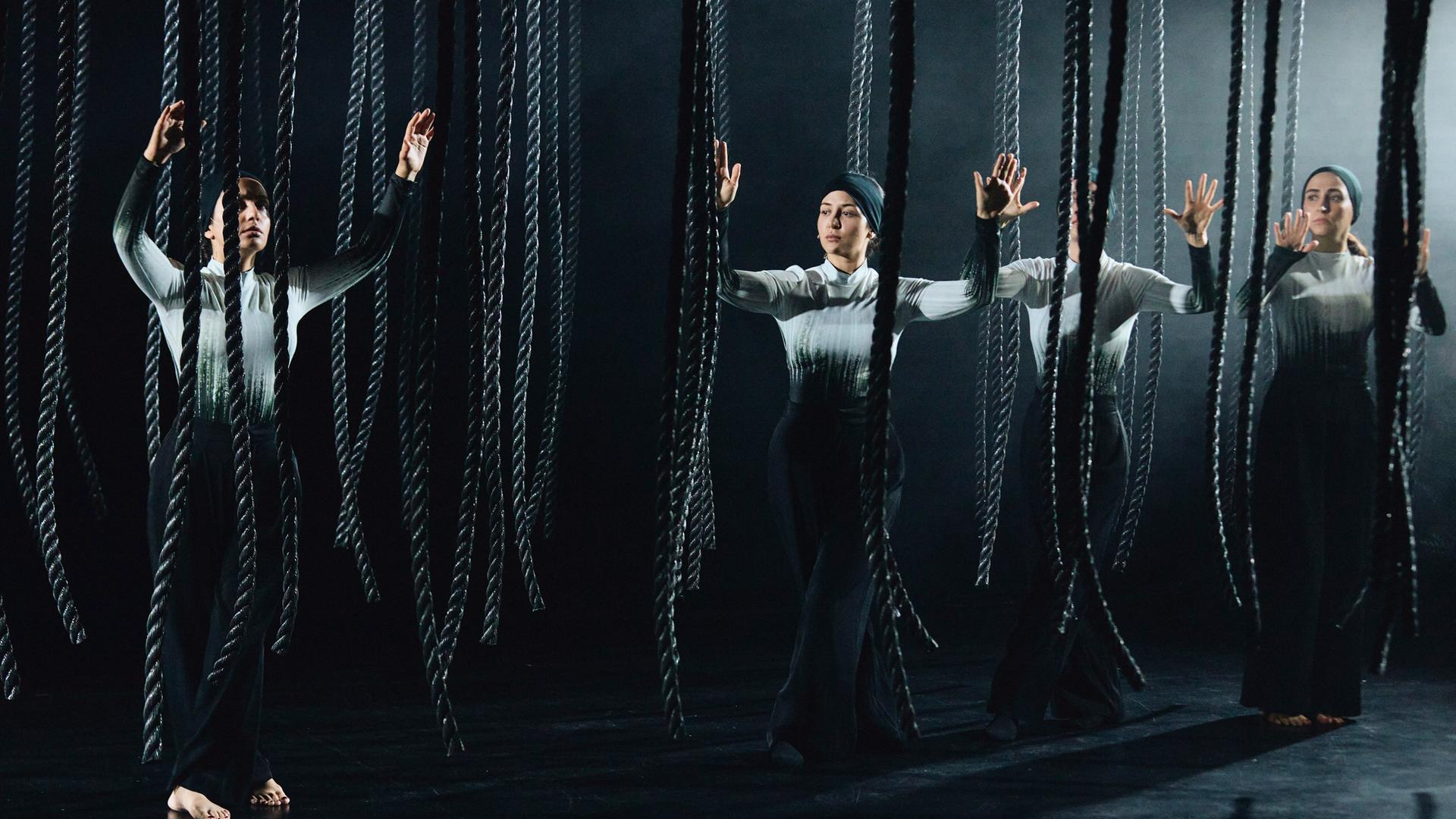 Bühnenszene: Frauen stehen mit erhobenen Händen zwischen herabhängenden Seilen.