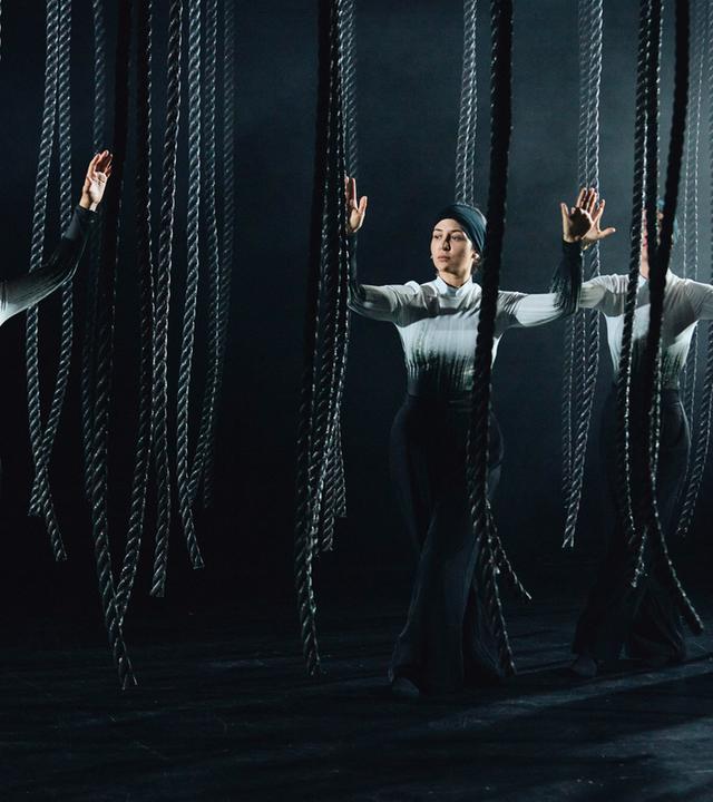 Bühnenszene: Frauen stehen mit erhobenen Händen zwischen herabhängenden Seilen.