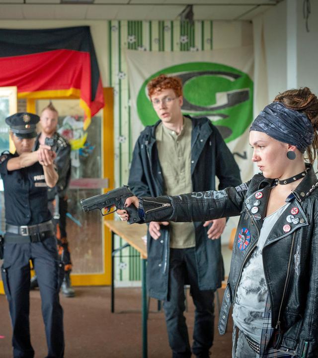"Notruf Hafenkante - Rattenburg": In einem mit Sprüchen, Plakaten und der Deutschlandfahne behängten Raum steht die linksautonome Ira Kaczinski (Emma Drogunova) Leo (Max Befort) gegenüber. Sie hat eine Waffe auf seinen Kopf gerichtet. Beide sehen sich in die Augen. Im Hintergrund stehen die Polizisten Hans Moor (Bruno F. Apitz) und Franzi Jung (Rhea Harder-Vennewald) nebeneinander. Beide haben ihre Waffe auf Ira gerichtet. Neben ihnen steht Sebastian Voigt (Julius Feldmeier), der versucht auf Ira einzureden.