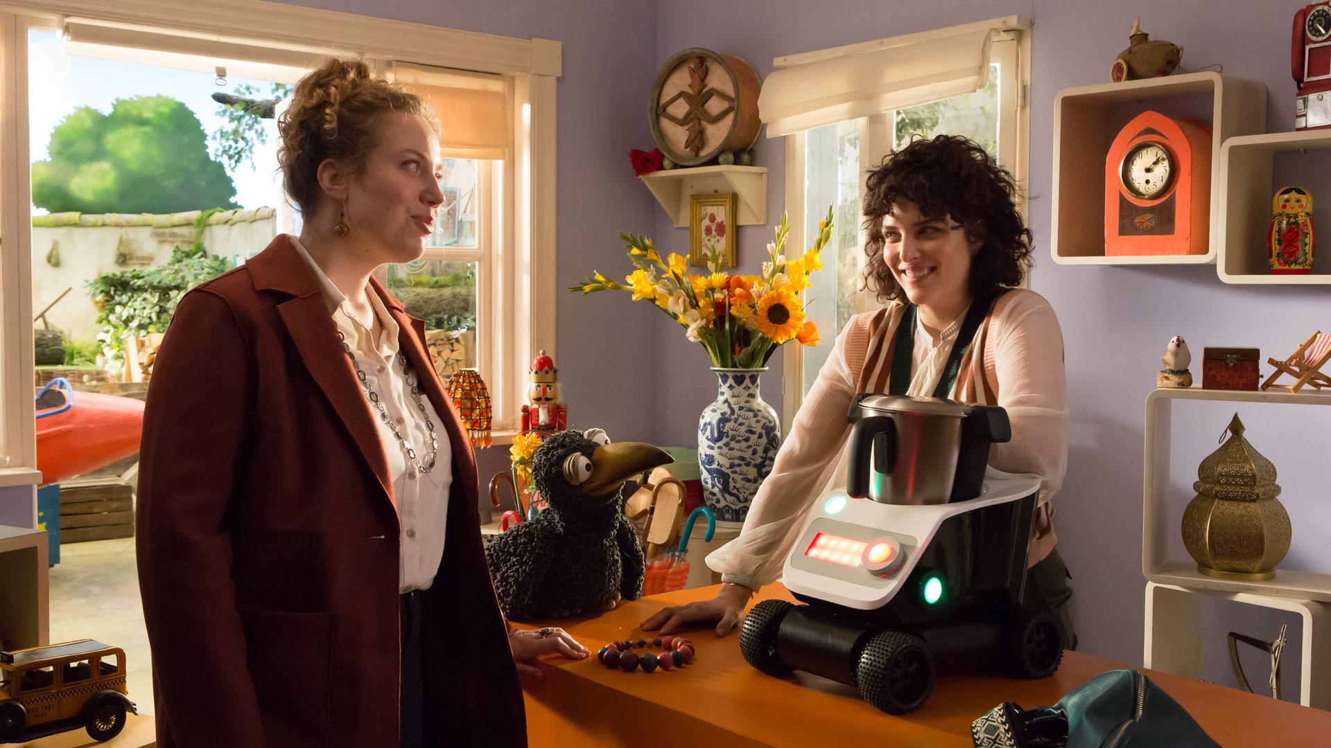 Frau Siebenstein steht im Laden hinter der Theke und schaut freundlich lächelnd zu einer Kundin. Auf der Theke steht eine große Küchenmaschine auf Rollen. Rudi sitzt daneben und schaut zu Frau Siebenstein. Im Hintergrund sieht man eine orange Uhr im Regal, einen Nussknacker, ein altes Auto und einen großen Blumenstrauß in einer großen blau-weißen Vase.