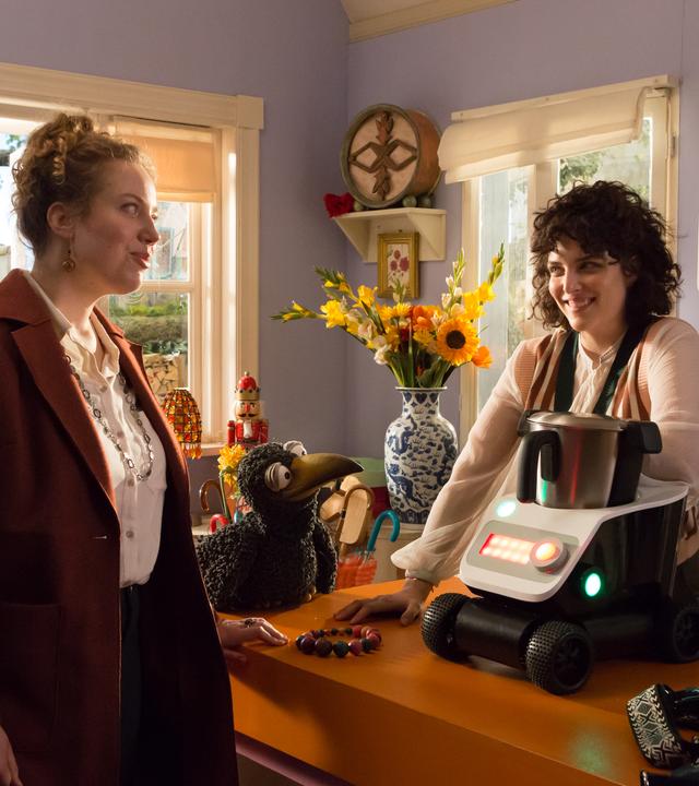 Frau Siebenstein steht im Laden hinter der Theke und schaut freundlich lächelnd zu einer Kundin. Auf der Theke steht eine große Küchenmaschine auf Rollen. Rudi sitzt daneben und schaut zu Frau Siebenstein. Im Hintergrund sieht man eine orange Uhr im Regal, einen Nussknacker, ein altes Auto und einen großen Blumenstrauß in einer großen blau-weißen Vase.