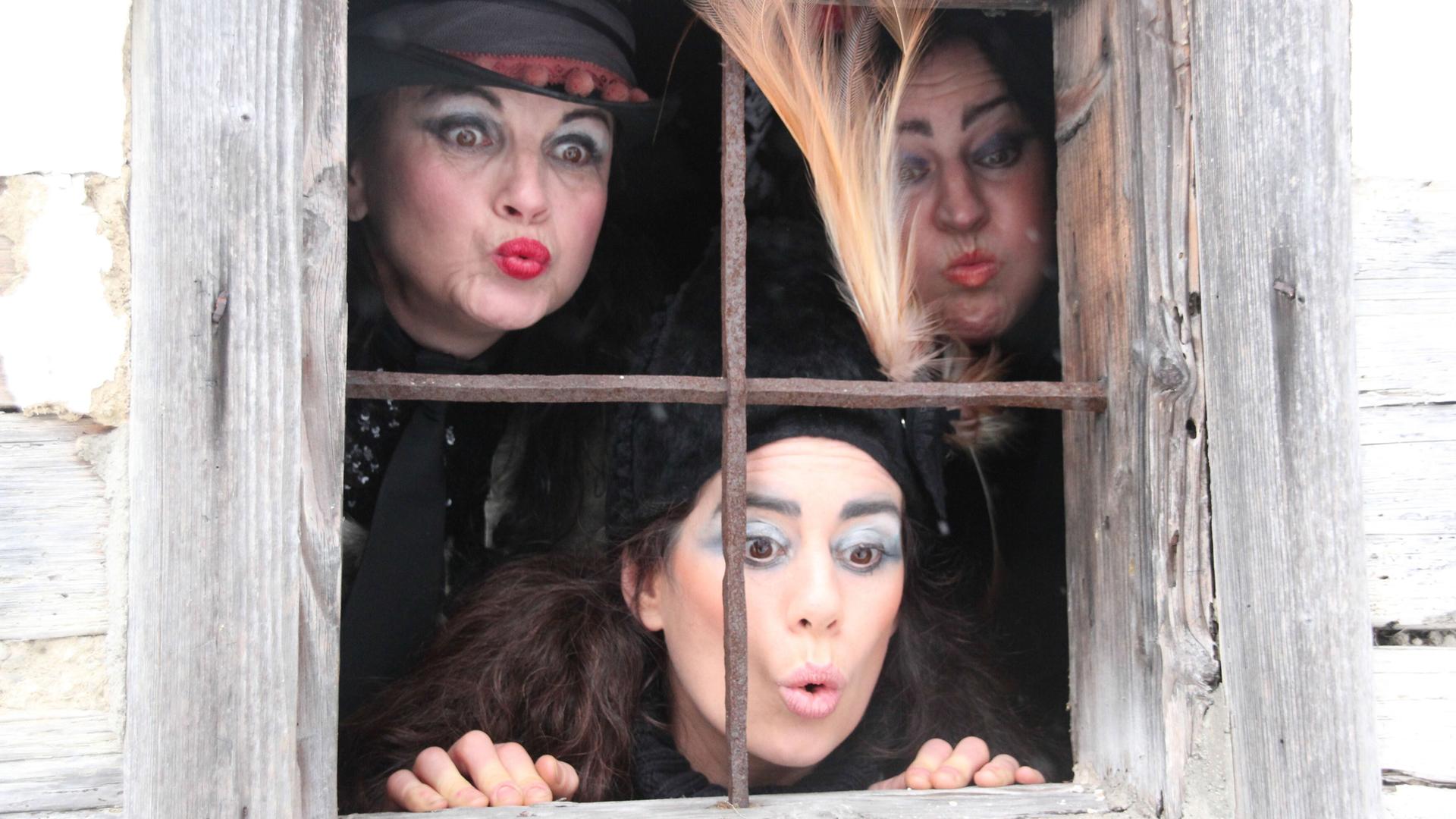 Das Bild zeigt eine Gruppe von drei Frauen, die durch ein Fenster schauen. Die Frauen tragen auffällige Kostüme, die an Hexen erinnern. Eine der Frauen hat einen schwarzen Hut mit einer Feder und eine lebhafte Schminkgestaltung, einschließlich auffälliger Augenfarbe und roten Lippen. Die anderen beiden Frauen haben ebenfalls markante Make-up-Styles, während sie verschiedene Gesichtsausdrücke zeigen, die Neugier und Überraschung ausstrahlen.   Das Fenster hat ein Gitter aus rostigem Metall und ist in eine rustikale Holzwand eingelassen. Im Hintergrund ist eine dunkle Umgebung zu erkennen. Die Gesamtatmosphäre des Bildes ist geheimnisvoll und spiegelt die magischen und mythologischen Themen wider, die mit den Rauhnachtgeschichten verbunden sind.