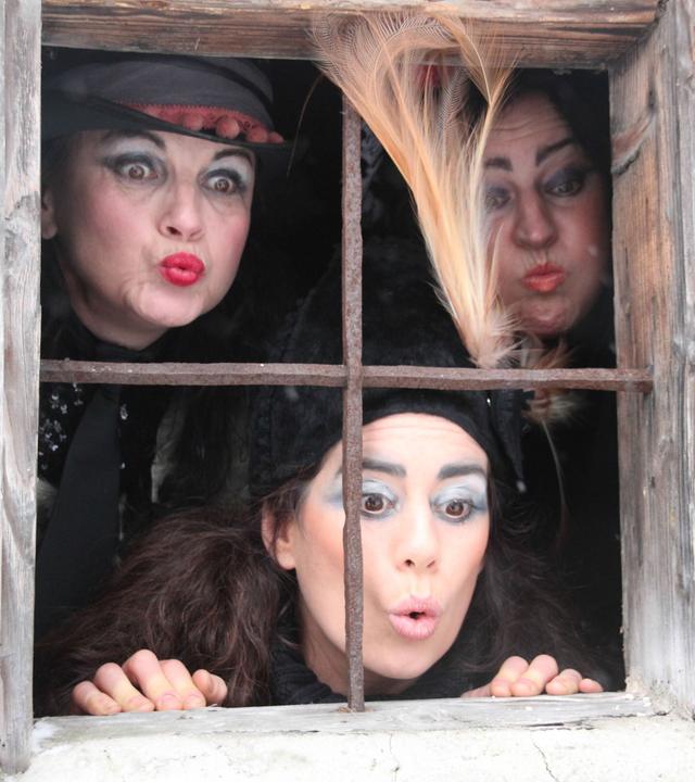 Das Bild zeigt eine Gruppe von drei Frauen, die durch ein Fenster schauen. Die Frauen tragen auffällige Kostüme, die an Hexen erinnern. Eine der Frauen hat einen schwarzen Hut mit einer Feder und eine lebhafte Schminkgestaltung, einschließlich auffälliger Augenfarbe und roten Lippen. Die anderen beiden Frauen haben ebenfalls markante Make-up-Styles, während sie verschiedene Gesichtsausdrücke zeigen, die Neugier und Überraschung ausstrahlen.   Das Fenster hat ein Gitter aus rostigem Metall und ist in eine rustikale Holzwand eingelassen. Im Hintergrund ist eine dunkle Umgebung zu erkennen. Die Gesamtatmosphäre des Bildes ist geheimnisvoll und spiegelt die magischen und mythologischen Themen wider, die mit den Rauhnachtgeschichten verbunden sind.