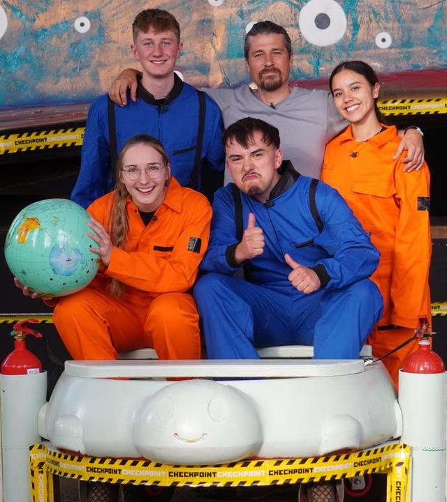 Checkpoint: Die Testteamer sitzen und stehen mit orangenen und blauen "Raumfahrtanzügen" in der Checkpoint-Halle, Laura hält einen Globus in der Hand. Tommy Scheel steht ebenfalls in der Gruppe.
