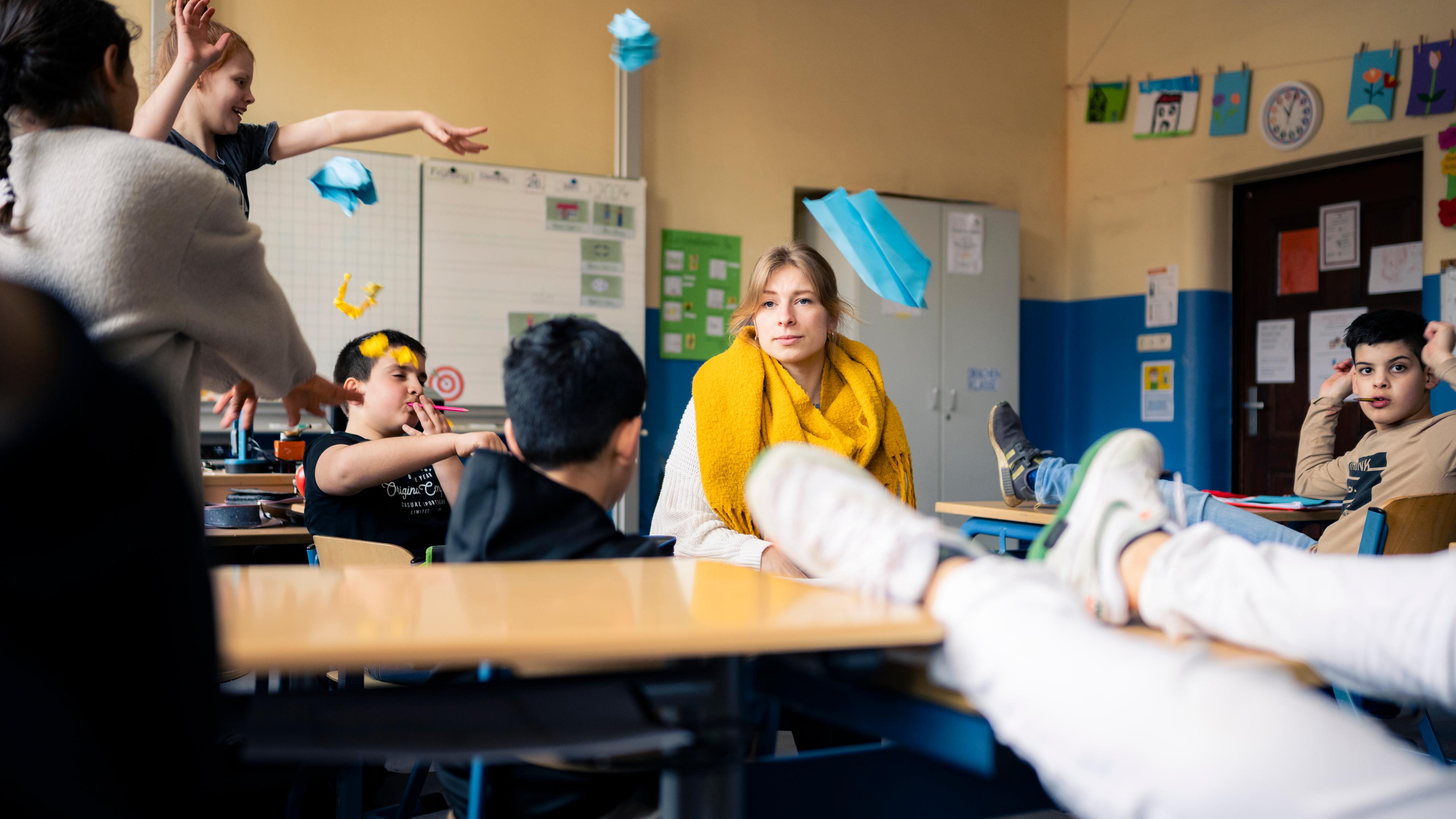 Lara sitzt inmitten einer Schulklasse. Schülerinnen und Schüler haben die Füße auf den Tischen und werfen Papierflieger durch den Klassenraum.