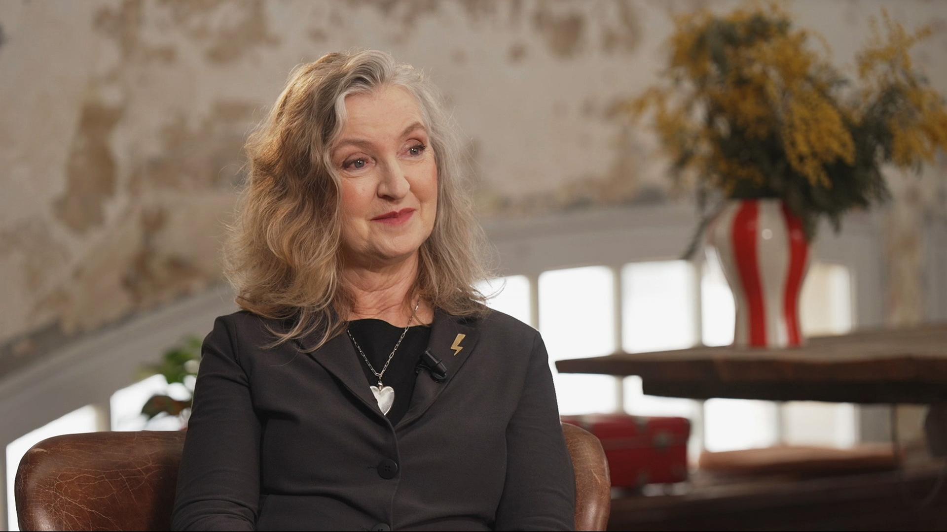 Rebecca Solnit sitzt in einem Sessel und lächelt leicht, trägt einen schwarzen Blazer und ein Herzcollier; im Hintergrund unscharf eine Vase mit gelben Blüten vor einer hellen Wand.