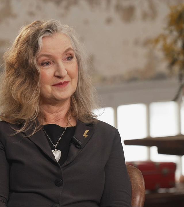 Rebecca Solnit sitzt in einem Sessel und lächelt leicht, trägt einen schwarzen Blazer und ein Herzcollier; im Hintergrund unscharf eine Vase mit gelben Blüten vor einer hellen Wand.