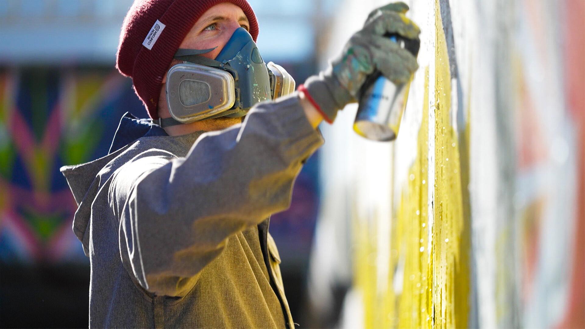 Das Bild zeigt einen männlichen Graffiti-Künstler, der an einer Wand arbeitet. Er trägt eine graue Jacke und eine rote Mütze. Sein Gesicht ist teilweise mit einer Atemschutzmaske bedeckt. In seiner rechten Hand hält er eine Sprühdose, mit der er gelbe Farbe auf die Wand sprüht. Im Hintergrund sind unscharfe farbige Muster zu erkennen, die vermutlich von vorherigen Graffiti stammen. Der Künstler scheint konzentriert zu sein und die kreative Aktivität zu genießen. Die Szene vermittelt eine Atmosphäre von urbaner Kunst und Ausdruck.