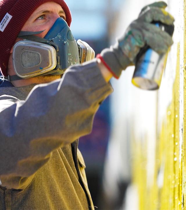 Das Bild zeigt einen männlichen Graffiti-Künstler, der an einer Wand arbeitet. Er trägt eine graue Jacke und eine rote Mütze. Sein Gesicht ist teilweise mit einer Atemschutzmaske bedeckt. In seiner rechten Hand hält er eine Sprühdose, mit der er gelbe Farbe auf die Wand sprüht. Im Hintergrund sind unscharfe farbige Muster zu erkennen, die vermutlich von vorherigen Graffiti stammen. Der Künstler scheint konzentriert zu sein und die kreative Aktivität zu genießen. Die Szene vermittelt eine Atmosphäre von urbaner Kunst und Ausdruck.