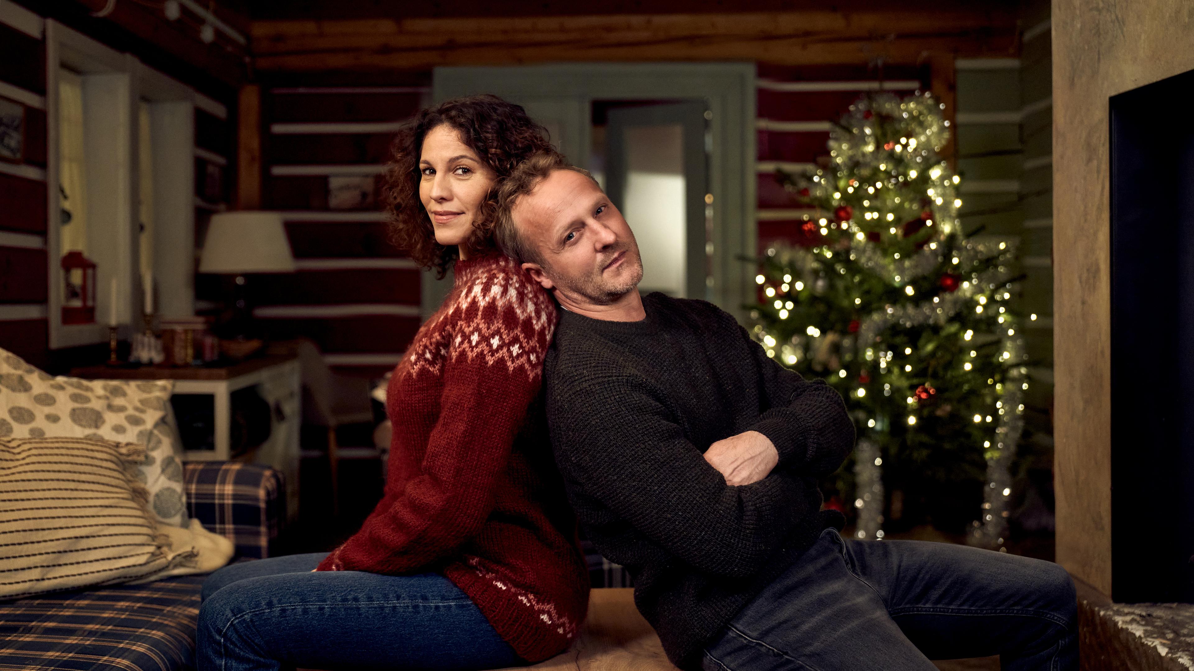 "Ein Regenbogen zu Weihnachten": In einer  gemütlich eingerichteten Skihütte sitzen Nicole Golding (Jasmin Gerat) und Martin Kupfer (Maximilian Brückner) Rücken an Rücken und schauen lächelnd in die Kamera. Im Hintergrund steht ein festlich geschmückter Weihnachtsbaum.