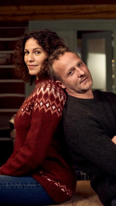 "Ein Regenbogen zu Weihnachten": In einer  gemütlich eingerichteten Skihütte sitzen Nicole Golding (Jasmin Gerat) und Martin Kupfer (Maximilian Brückner) Rücken an Rücken und schauen lächelnd in die Kamera. Im Hintergrund steht ein festlich geschmückter Weihnachtsbaum.