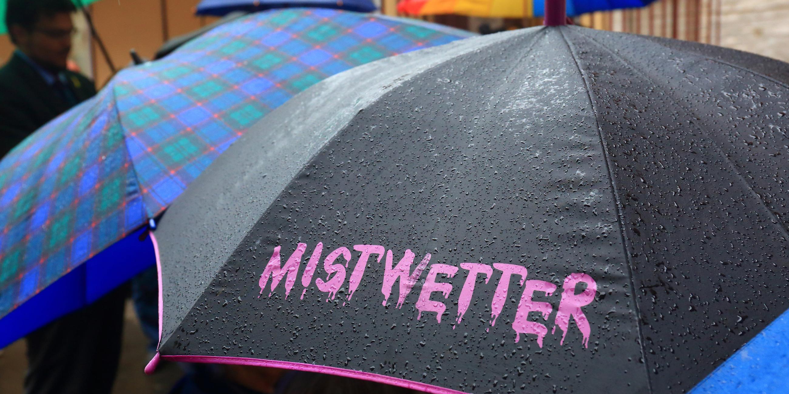 Zu sehen sind mehrere Regenschirme. Im Vordergrund ist ein schwarzer Regenschirm mit einer rosa Beschriftung: "Mistwetter".