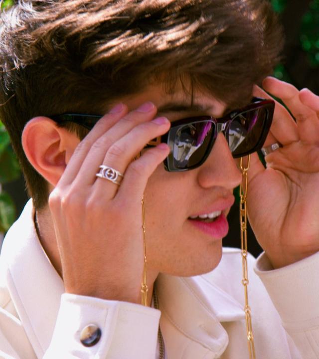 Influencer Oscar, junger Mann, ist weiß gekleidet und fasst mit seinen Händen seine Designer-Sonnenbrille.