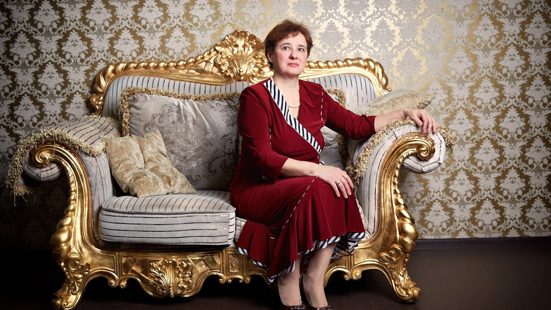 Eine Frau in einem roten Kleid sitzt steif und aufrecht auf einem vergoldeten Sofa vor einer mit goldener Brokattapete