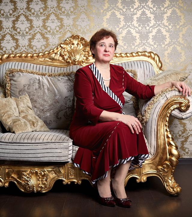 Eine Frau in einem roten Kleid sitzt steif und aufrecht auf einem vergoldeten Sofa vor einer mit goldener Brokattapete