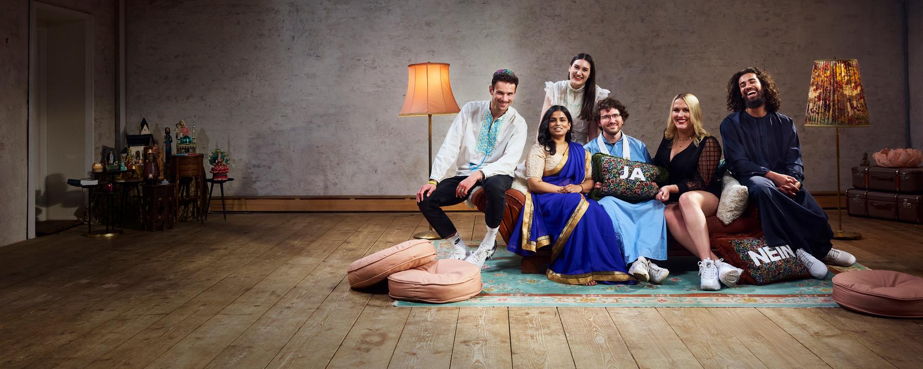Die sechs Bewohner der Glaubens-WG sitzen auf dem Sofa im Wohnzimmer und lachen gemeinsam in die Kamera.