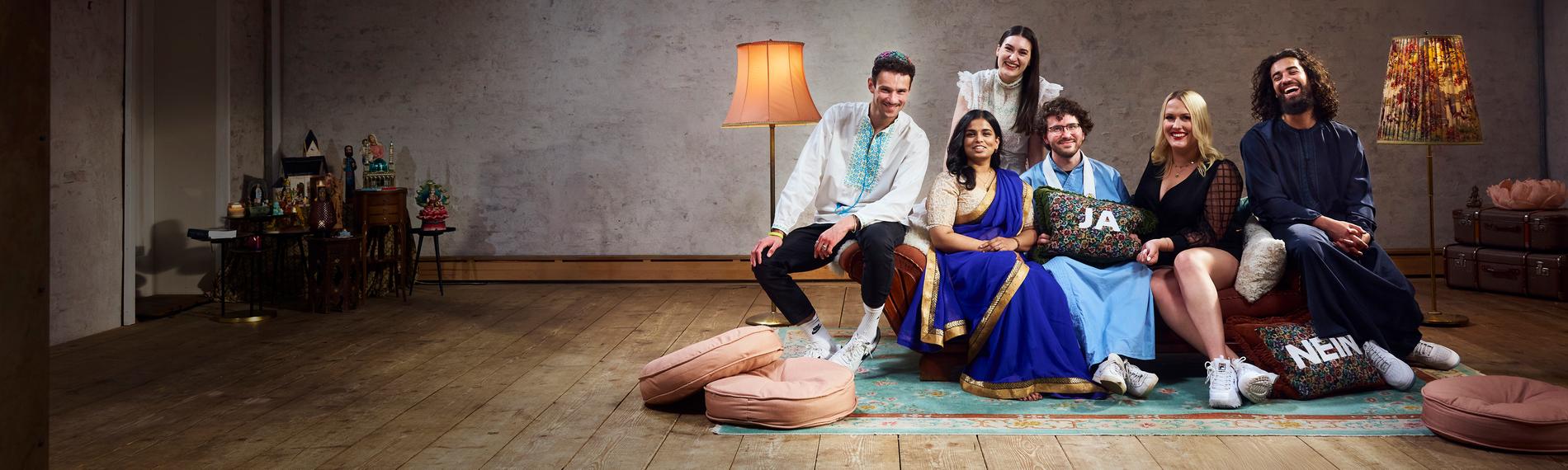 Die sechs Bewohner der Glaubens-WG sitzen auf dem Sofa im Wohnzimmer und lachen gemeinsam in die Kamera.