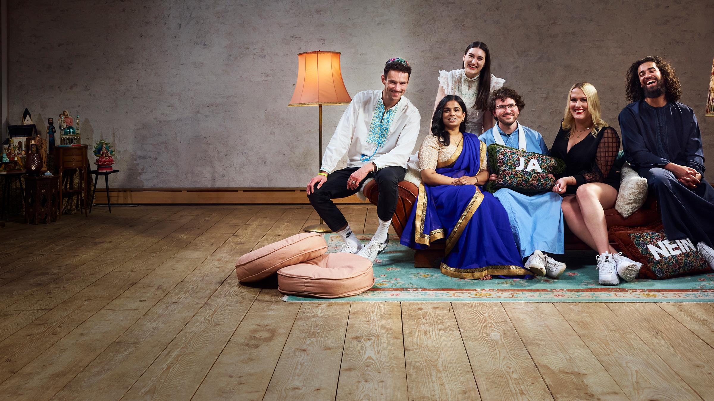 Die sechs Bewohner der Glaubens-WG sitzen auf dem Sofa im Wohnzimmer und lachen gemeinsam in die Kamera.