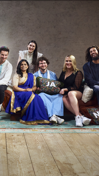 Die sechs Bewohner der Glaubens-WG sitzen auf dem Sofa im Wohnzimmer und lachen gemeinsam in die Kamera.