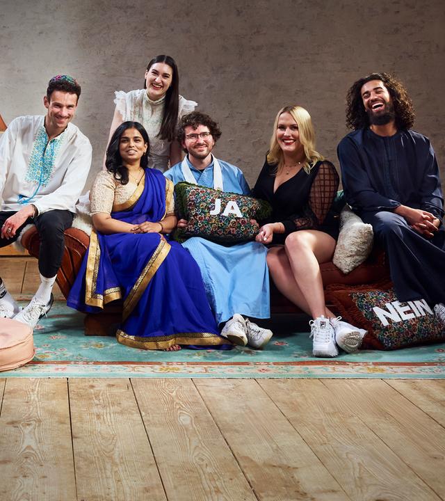 Die sechs Bewohner der Glaubens-WG sitzen auf dem Sofa im Wohnzimmer und lachen gemeinsam in die Kamera.