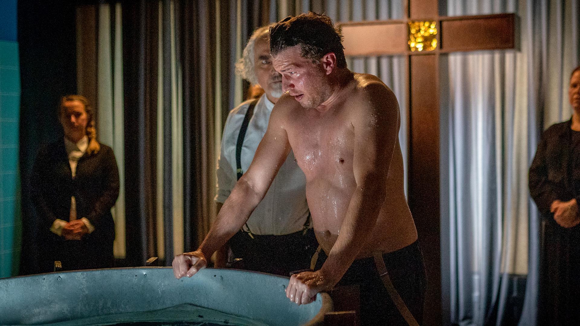 Nick Brandt (Raúl Richter) steht mit freiem Oberkörper in einem düsteren Raum. Er stützt sich mit beiden Händen an den Rand eines großen Bottiches, in dem Wasser ist. Sein Kopf und sein Oberkörper sind nass. Neben ihm steht Gideon Frey (Hansa Czypionka), der ihn ansieht. Im Hintergrund stehen zwei Frauen (Komparsen). Im Hintergrund steht ein großes Kreuz.