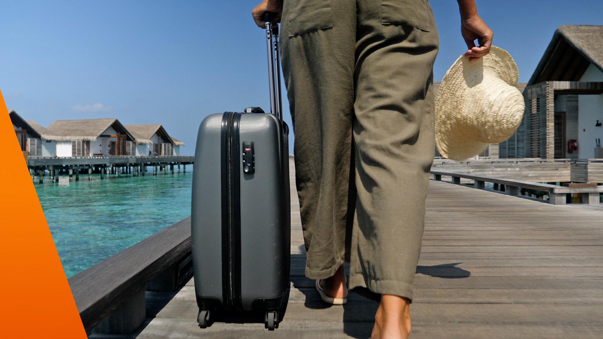 Frau läuft an einer Promenade lang, zieht ihren Koffer und hat einen Hut in der Hand