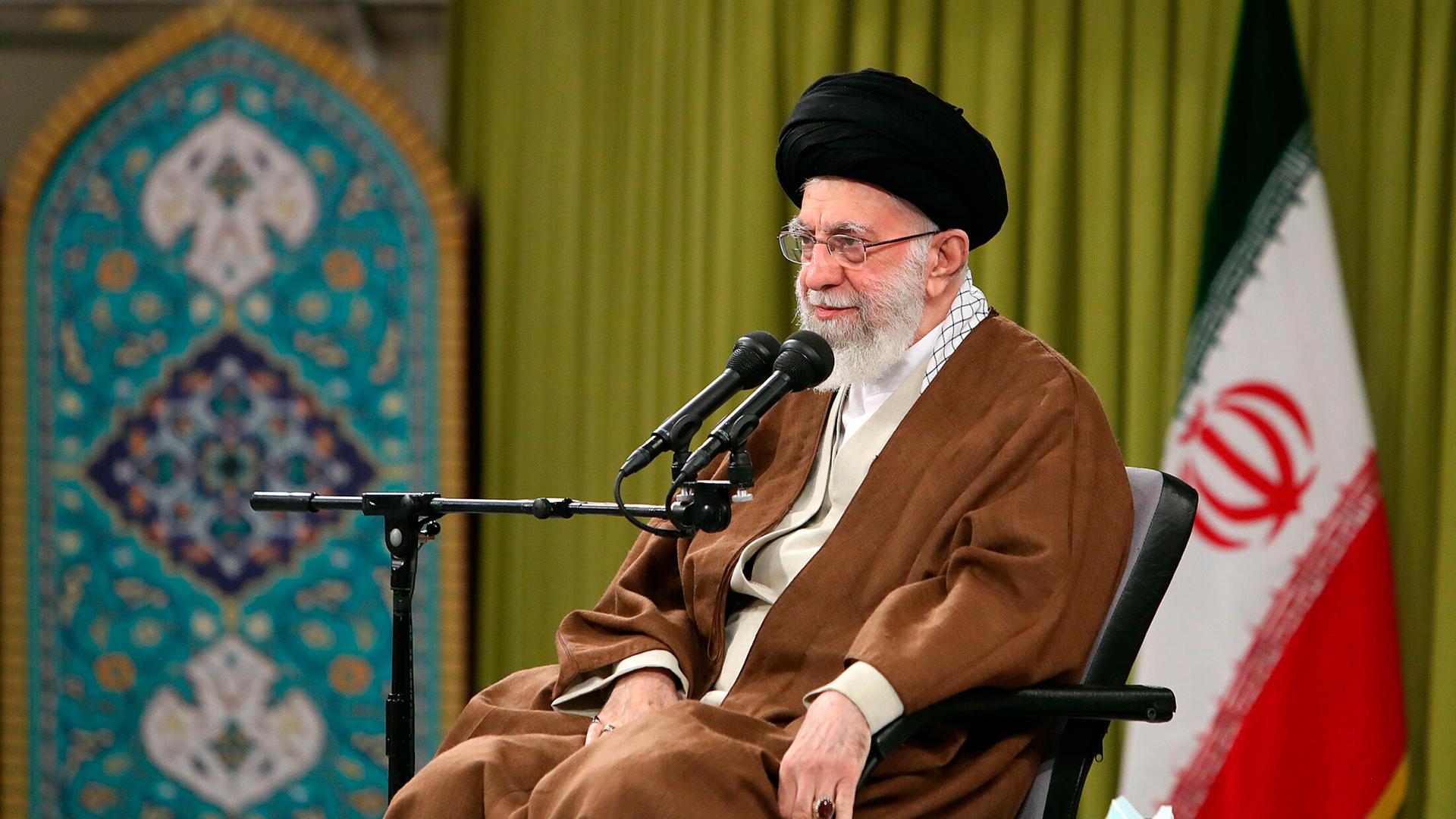 Ajatollah Ali Chamenei sitzt vor einem Mikrofon, im Hintergrund die iranische Flagge. 