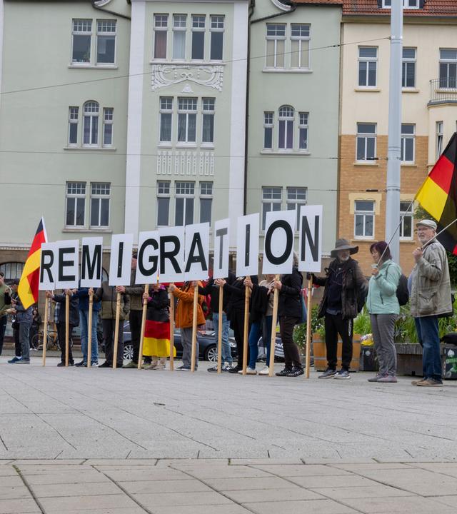 Frau mit Kopftuch geht an Demonstranten vorbei, die Buchstaben des Schriftzugs "Remigration" hochhalten.