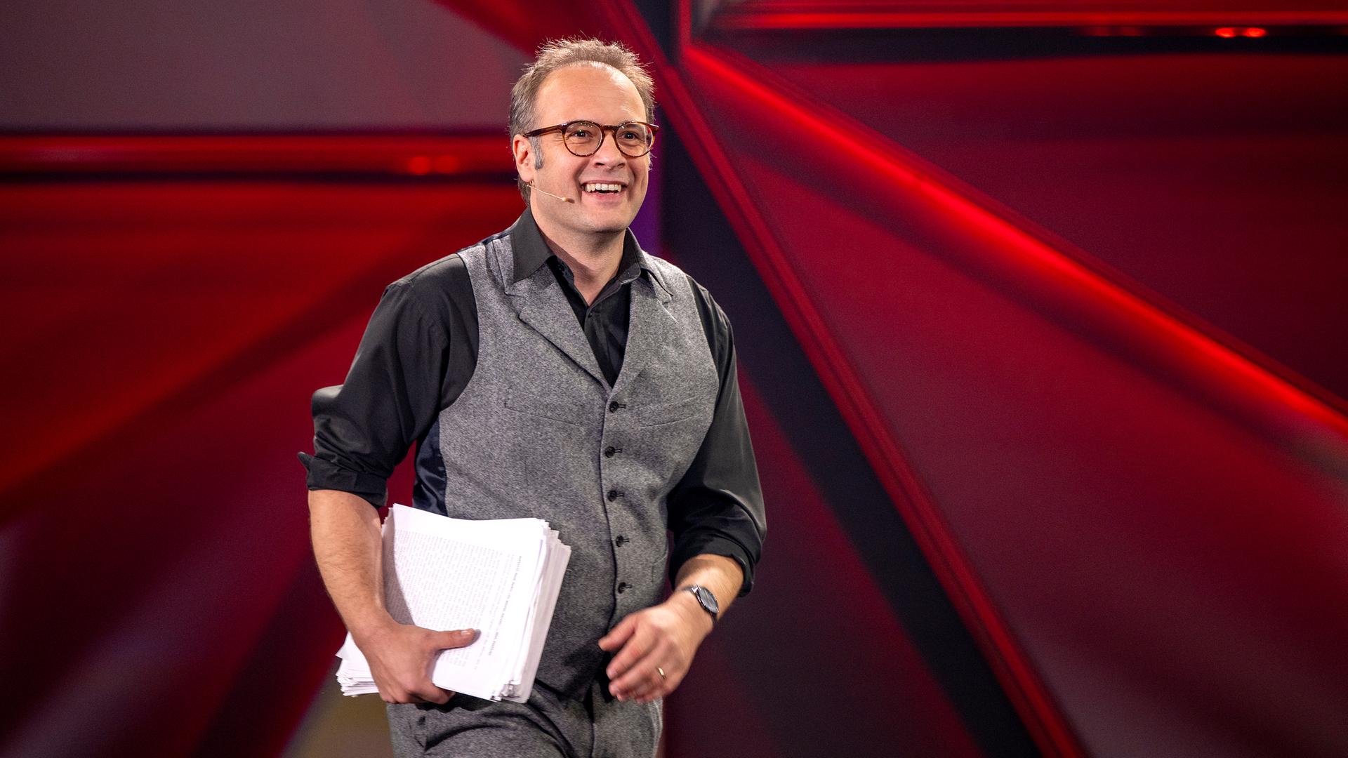 René Sydow ist in der Mitte des Bildes zu sehen. Er trägt einen Stapel Papier unter dem rechten Arm. Der Hintergrund ist rot beleuchtet.